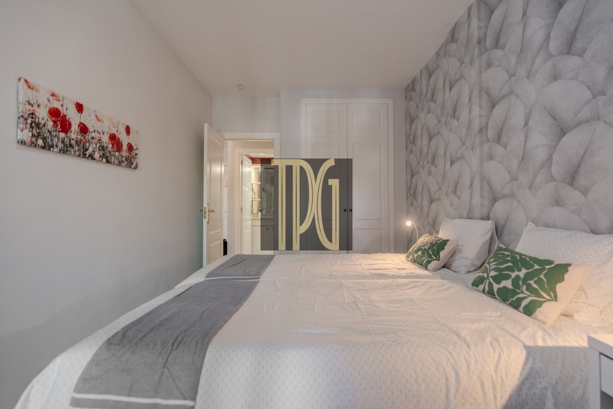 Hotel bedroom with a king-size bed, white linens, green leaf-pattern cushions, a grey textured wall, and a white wardrobe in the background.