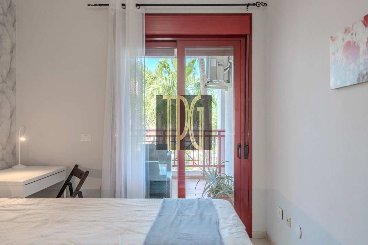 Bedroom with a red-framed glass door opening to a balcony, a white desk and lamp on the left.