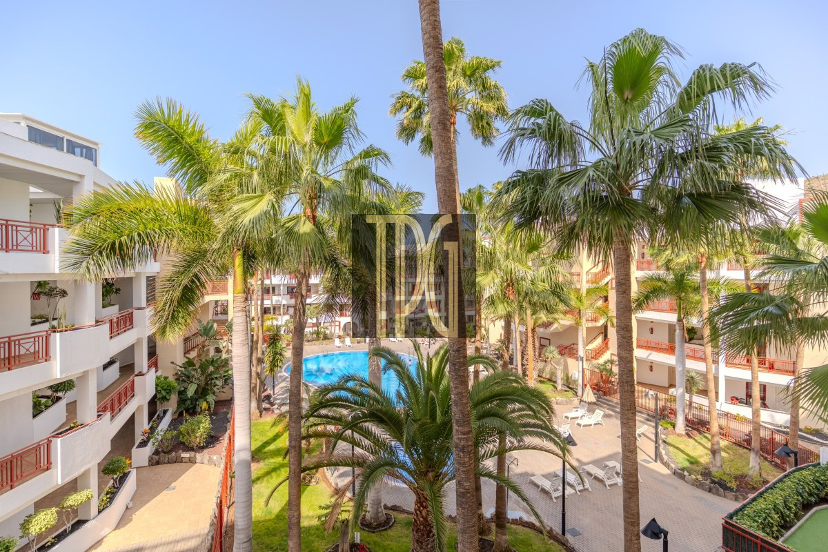 Resort courtyard with tall palm trees surrounding a blue swimming pool and white balconies of hotel rooms on either side.