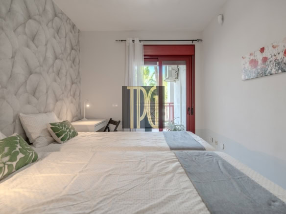 Twin-bed hotel-style room with a large quilted headboard, white bedding, and green leaf pillows; desk and lamp at the far end; red-framed glass door to balcony on the right; wall art with red flowers.