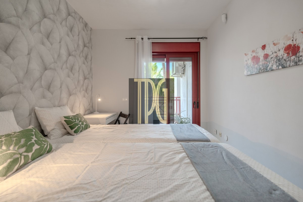 Twin-bed hotel-style room with a large quilted headboard, white bedding, and green leaf pillows; desk and lamp at the far end; red-framed glass door to balcony on the right; wall art with red flowers.