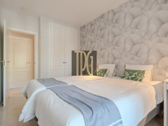 Bright hotel bedroom with a large bed, white bedding and gray throw, two green leaf-pattern cushions, white wardrobe, pale blue walls, and an open door on the left