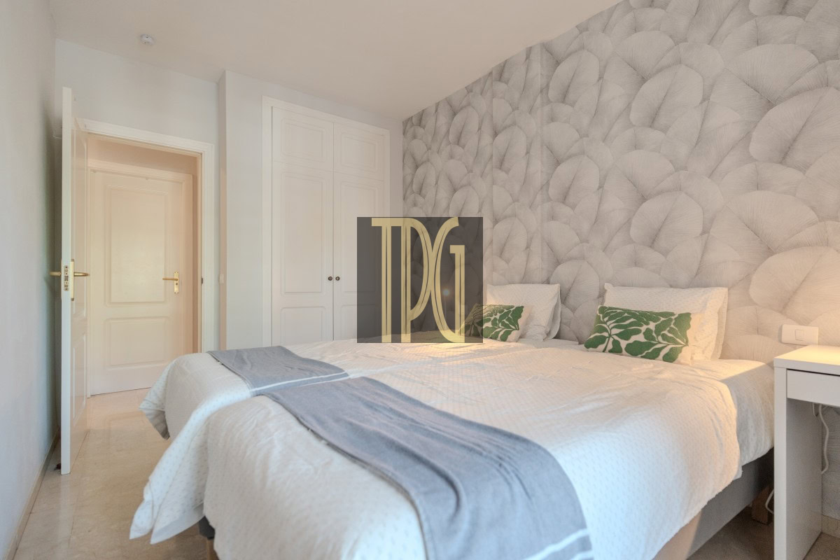 Bright hotel bedroom with a large bed, white bedding and gray throw, two green leaf-pattern cushions, white wardrobe, pale blue walls, and an open door on the left