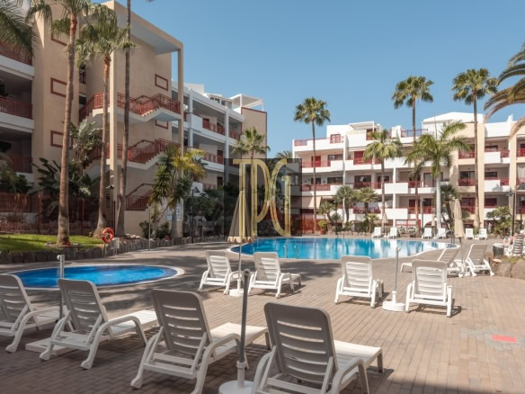 Resort pool area with white buildings, red railings, tall palm trees and rows of white lounge chairs around a large blue pool under a clear sky.