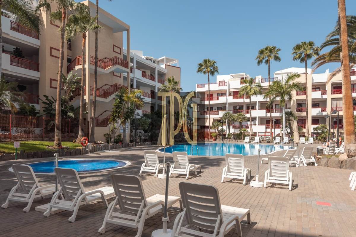 Resort pool area with white buildings, red railings, tall palm trees and rows of white lounge chairs around a large blue pool under a clear sky.