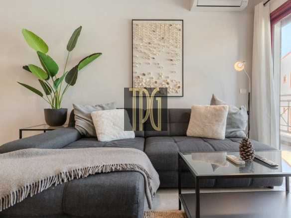 Cozy living room with a dark gray sectional sofa, plush pillows, and a light fringe throw, flanked by a potted plant and glass coffee table with a pine cone centerpiece.