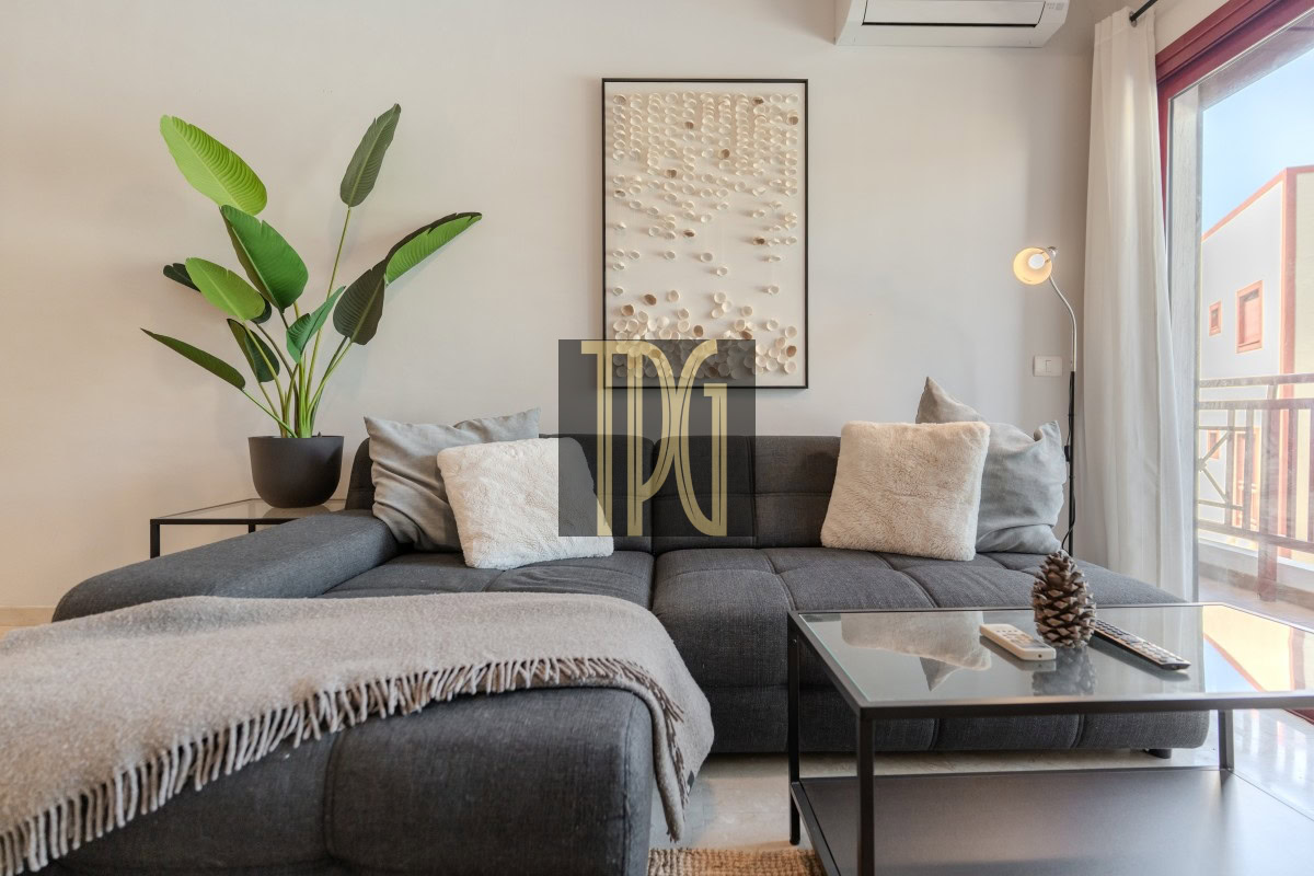 Cozy living room with a dark gray sectional sofa, plush pillows, and a light fringe throw, flanked by a potted plant and glass coffee table with a pine cone centerpiece.