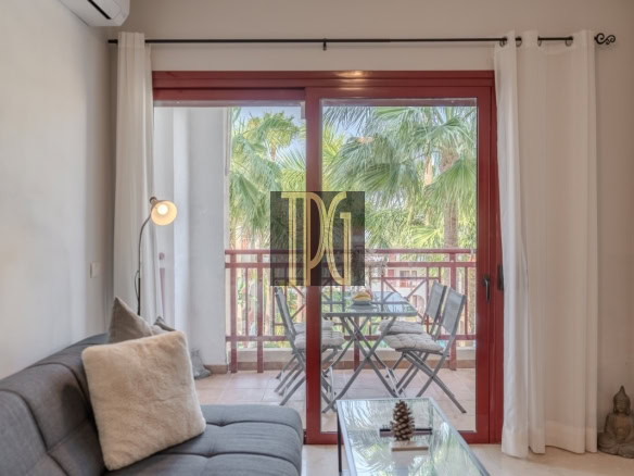 Living room with a gray sofa and beige throw pillow, facing a glass coffee table; red-framed sliding doors open to a balcony with a table and chairs and lush palm trees outside.