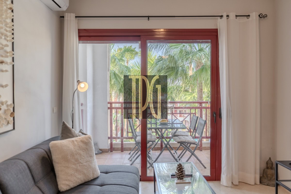 Living room with a gray sofa and beige throw pillow, facing a glass coffee table; red-framed sliding doors open to a balcony with a table and chairs and lush palm trees outside.