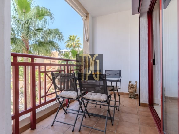 Outdoor balcony with a metal table and four folding chairs, red railing, and palm trees in the background
