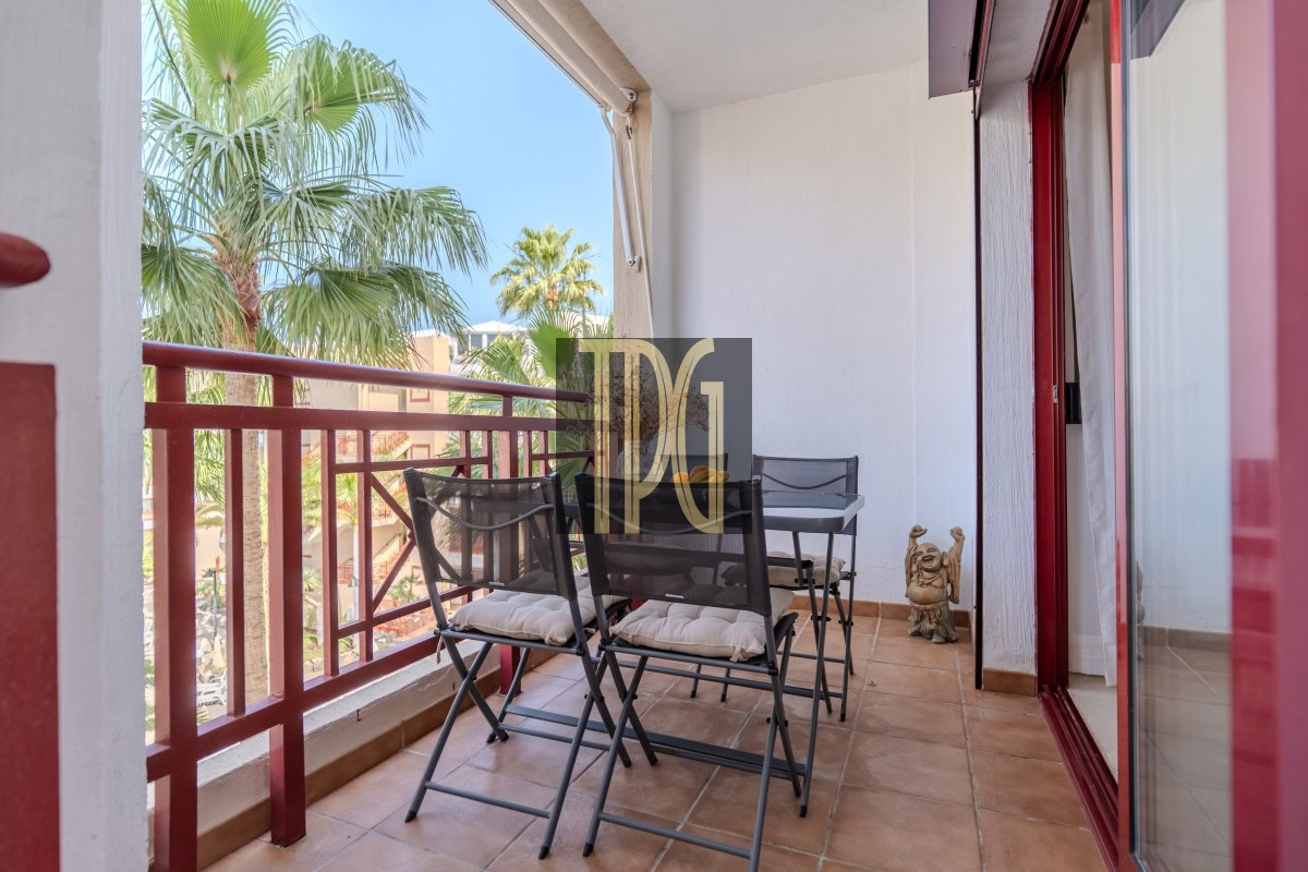 Outdoor balcony with a metal table and four folding chairs, red railing, and palm trees in the background