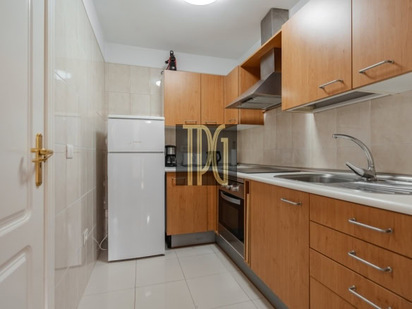 Compact galley kitchen with wooden cabinets, white fridge, oven, and stainless steel sink along one wall.