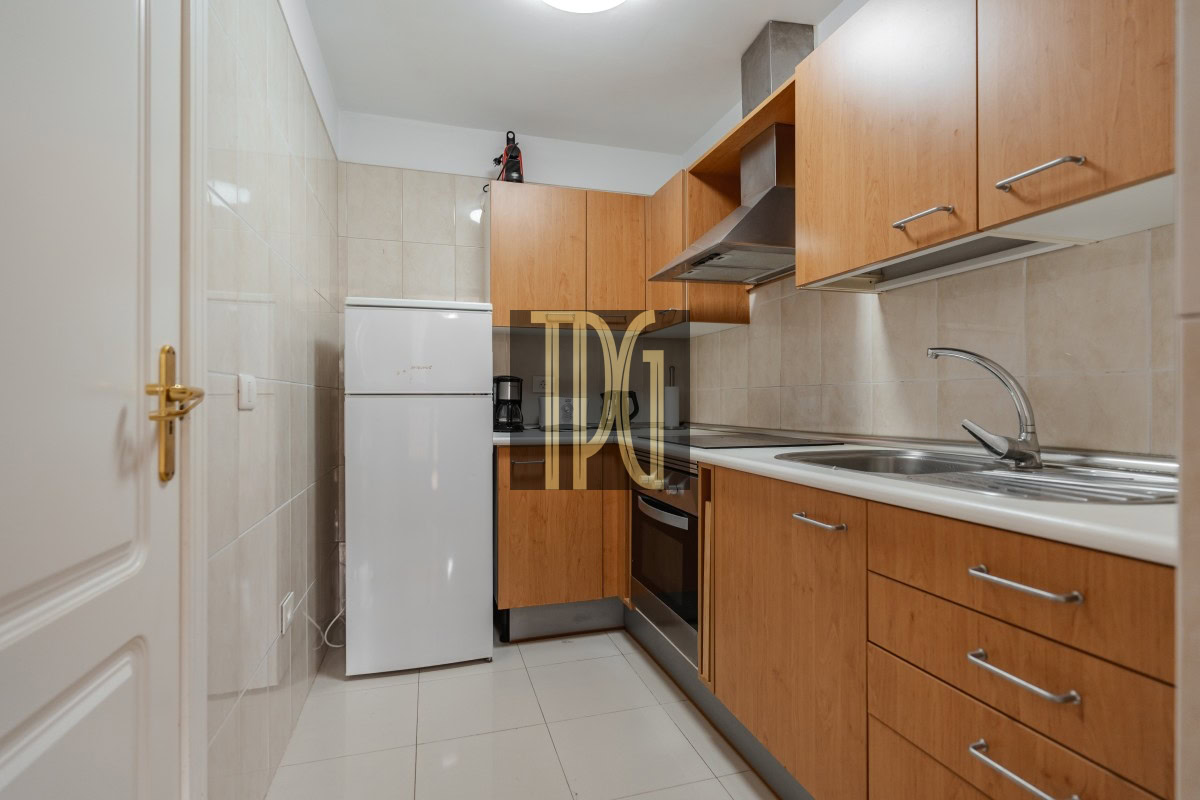 Compact galley kitchen with wooden cabinets, white fridge, oven, and stainless steel sink along one wall.