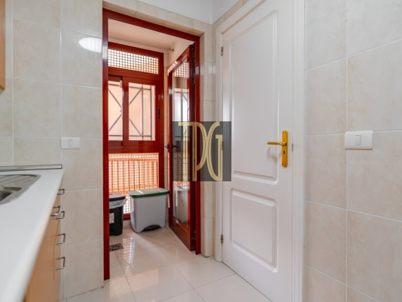 Kitchen with beige tiled walls, white cabinets and sink on the left, and a red-framed door opening to a balcony with a trash can outside outside the doorway.