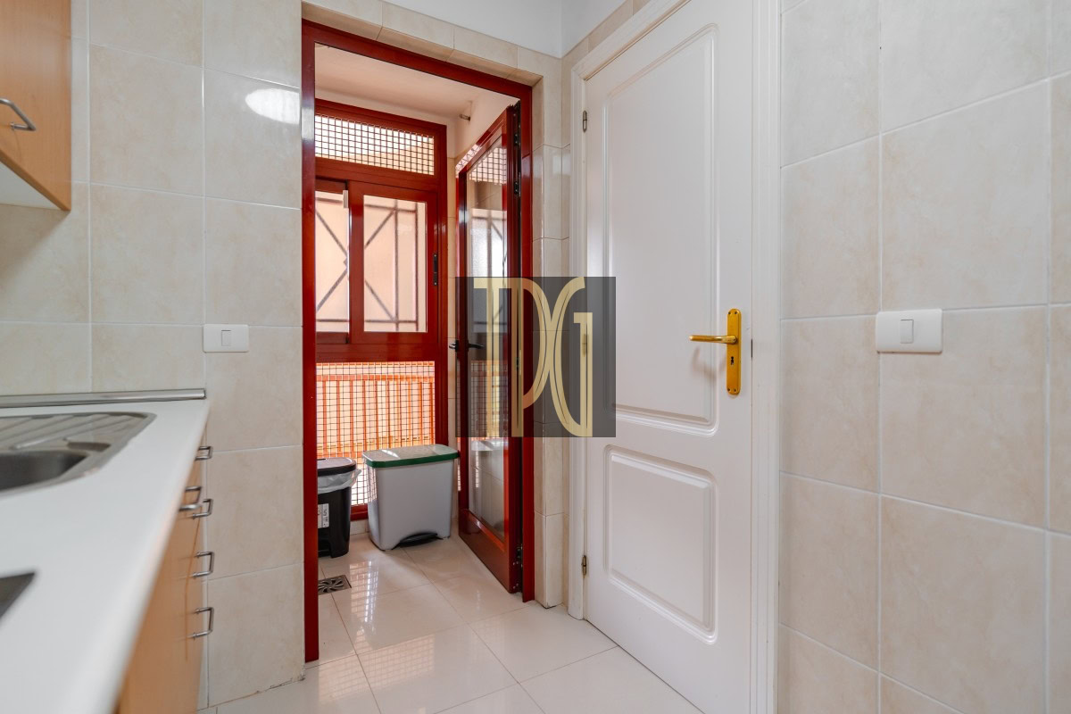 Kitchen with beige tiled walls, white cabinets and sink on the left, and a red-framed door opening to a balcony with a trash can outside outside the doorway.