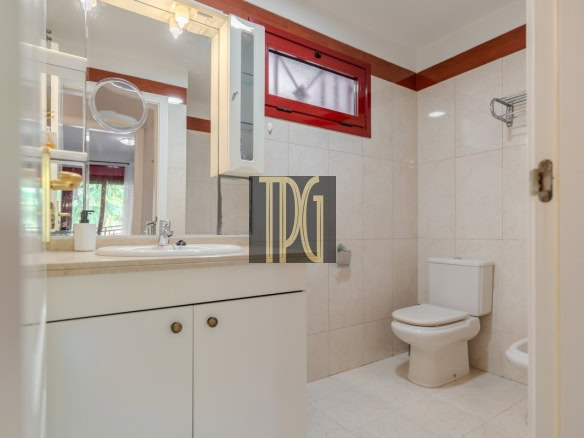 Bright bathroom with white vanity, sink, and cabinet; mirror above; white toilet and a red-framed window.