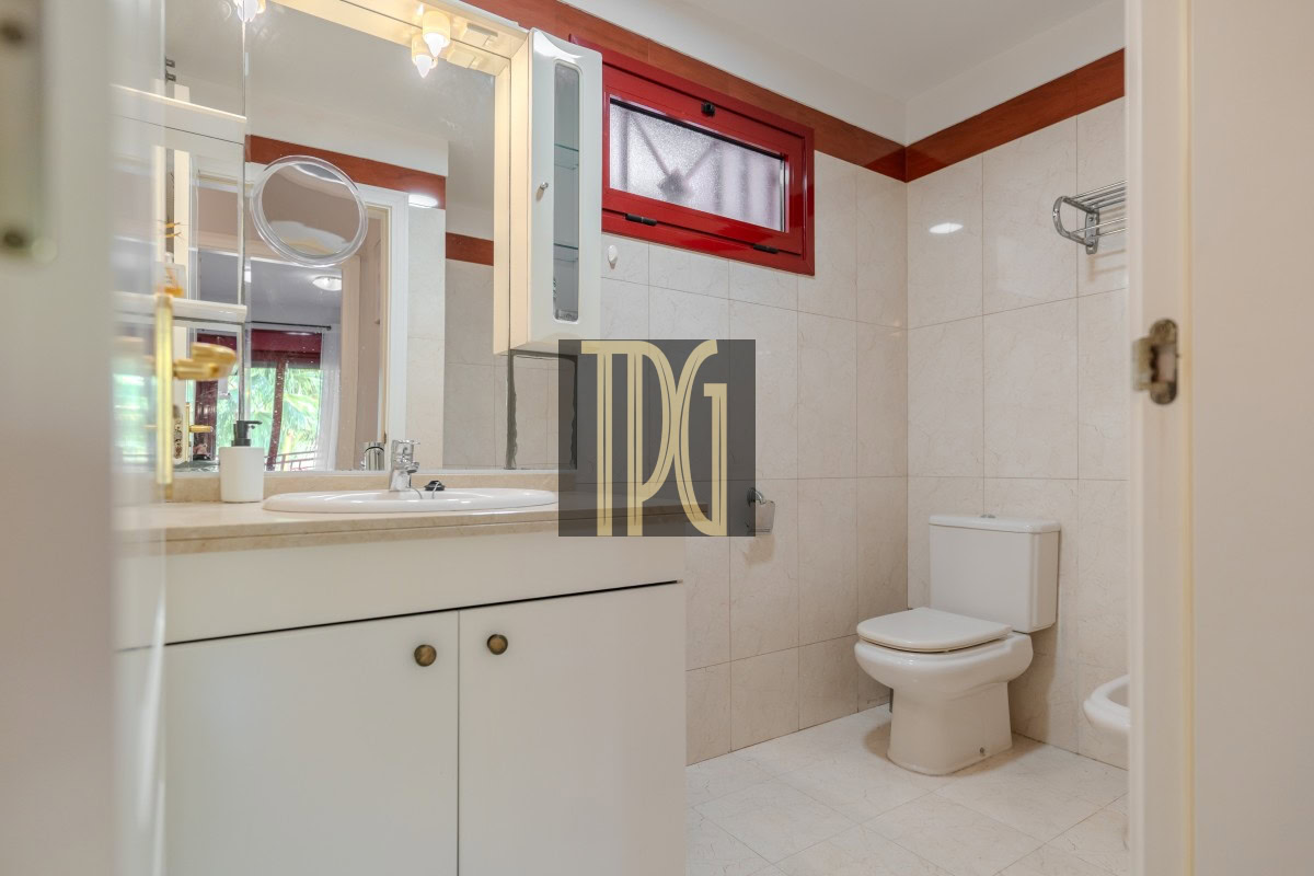 Bright bathroom with white vanity, sink, and cabinet; mirror above; white toilet and a red-framed window.