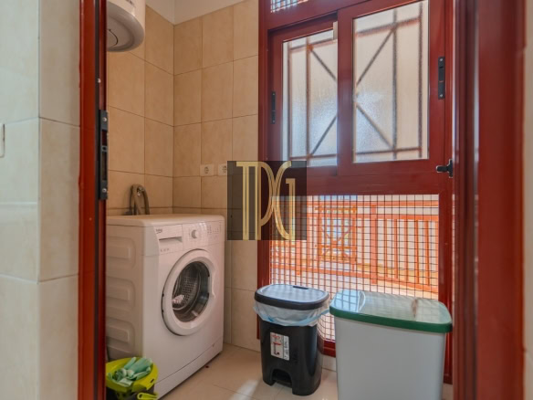 Small laundry nook with a front-loading washing machine beside tiled walls, a black trash bin with a blue bag, and a gray recycling bin by a red-framed door with frosted glass panels.