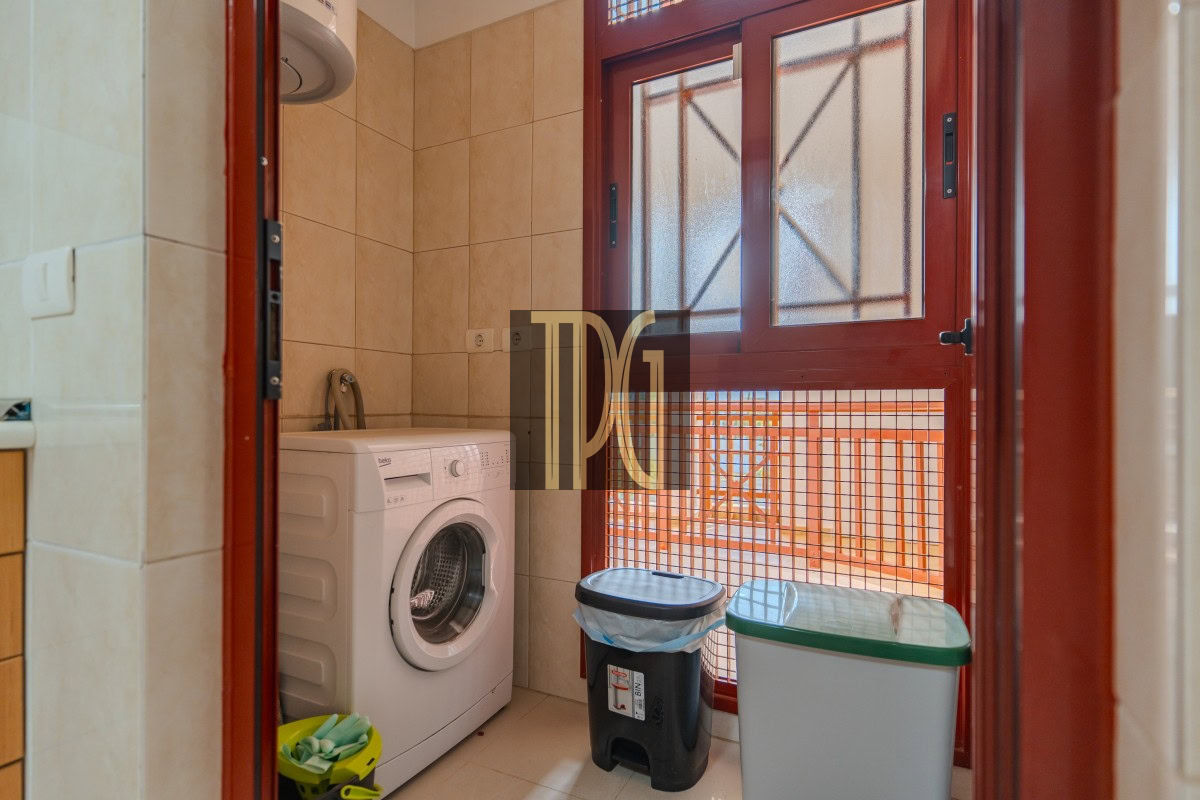 Small laundry nook with a front-loading washing machine beside tiled walls, a black trash bin with a blue bag, and a gray recycling bin by a red-framed door with frosted glass panels.