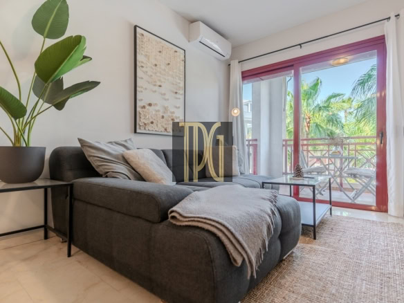 Modern living room with a gray sectional, beige throw, and a plant on a side table beside a wall art piece, opening to a sunny balcony through glass doors.