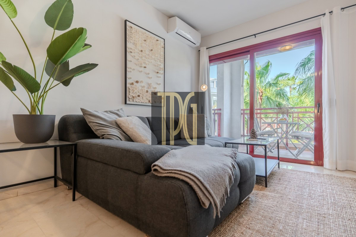 Modern living room with a gray sectional, beige throw, and a plant on a side table beside a wall art piece, opening to a sunny balcony through glass doors.