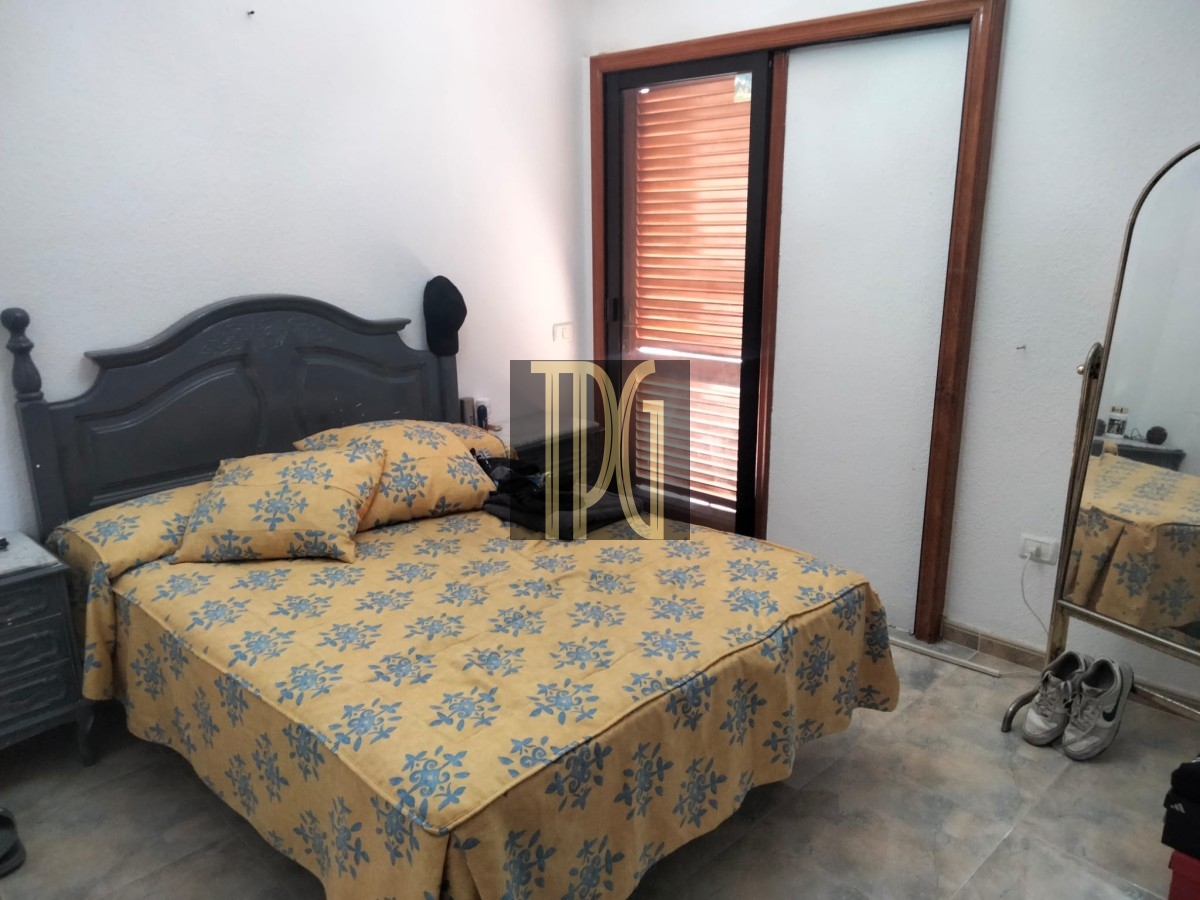 Bedroom with a gray ornate headboard, yellow floral bedspread, and matching pillows; a small nightstand on the left, and a full-length mirror with a wooden frame on the right. A door with wooden blinds is at the far wall, and sneakers sit on the floor near the mirror.