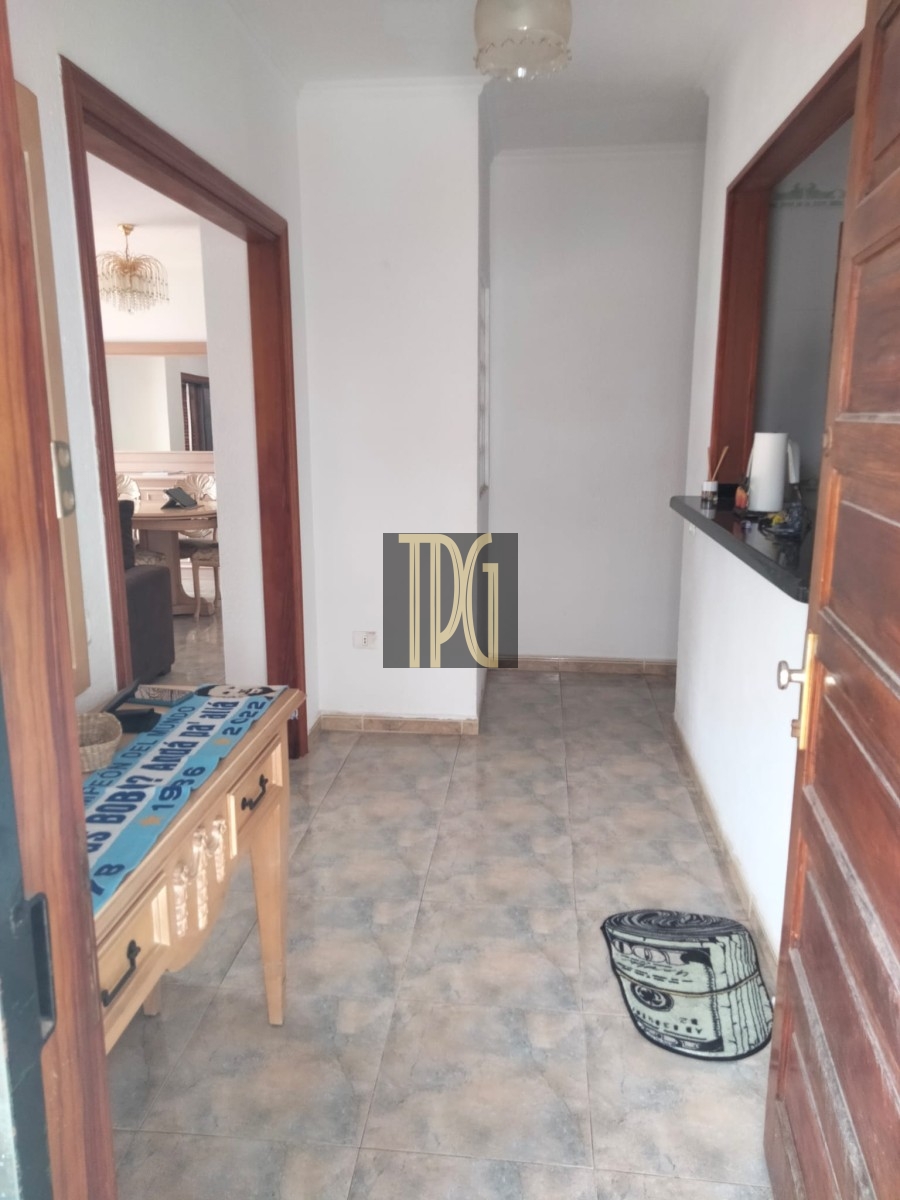 Narrow entry hall with gray tiled floor; left doorway opens to a living area with a chandelier, right side a kitchen counter with items on it, an open wooden door, a wooden console table with a blue patterned runner, and a round drum-shaped ottoman on the floor.