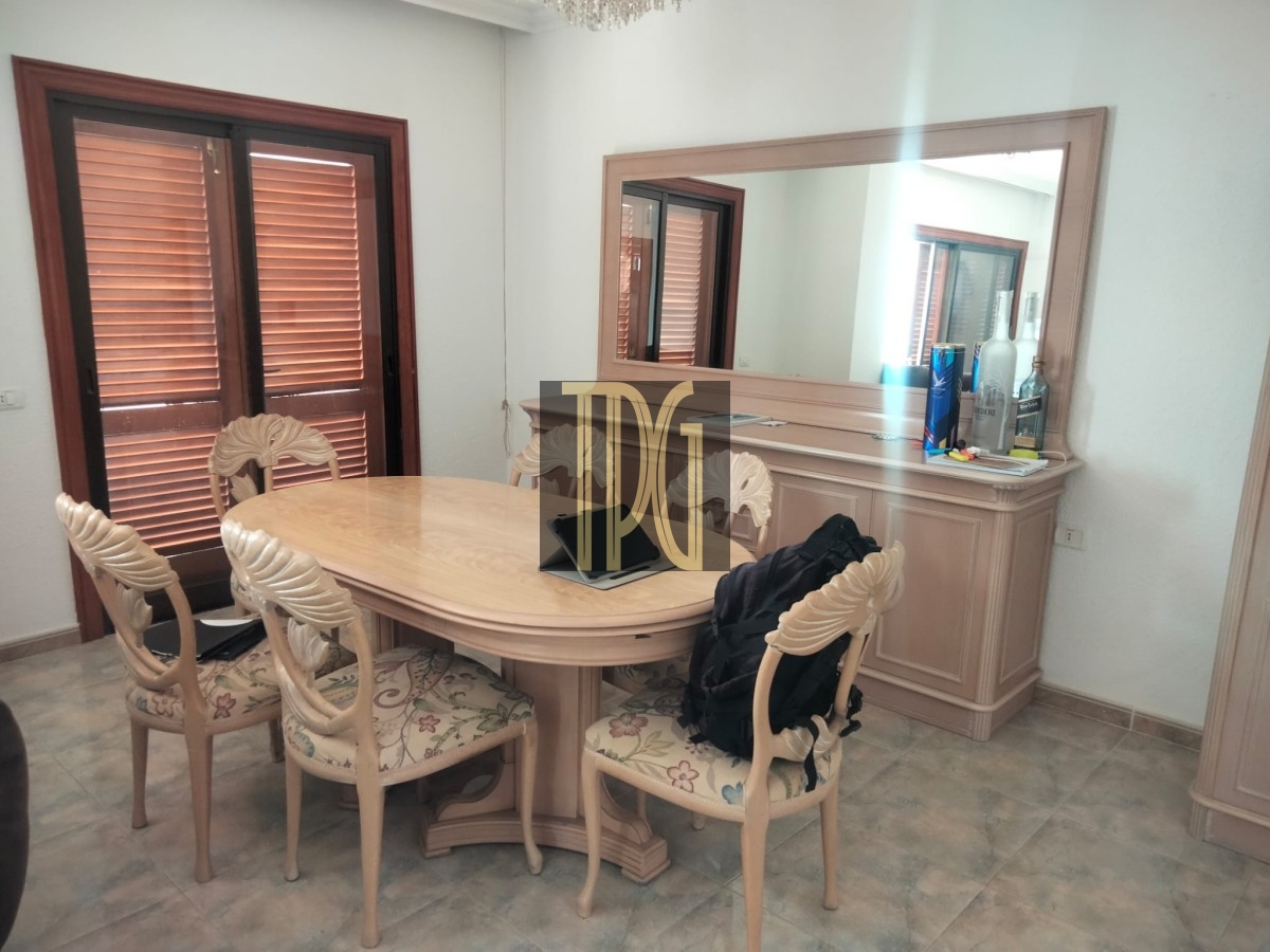 Cream-colored dining room with an oval table, carved chairs, and a large mirror above a sideboard; wooden shutters cover the windows, and a jacket rests on a chair beside a laptop on the table.