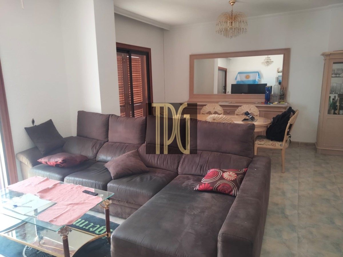 Living room with a dark brown sectional sofa, pink cushions, and a glass coffee table; dining area visible in the background with a large mirror and chandelier.