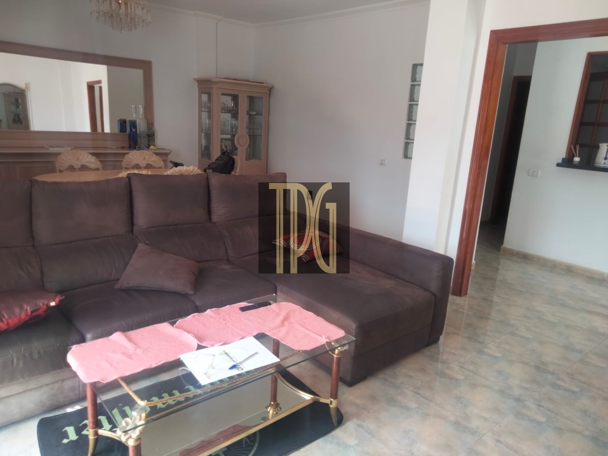 Brown L-shaped sofa in a living room with a glass coffee table and pink placemats, tiled floor, and a china cabinet in the background
