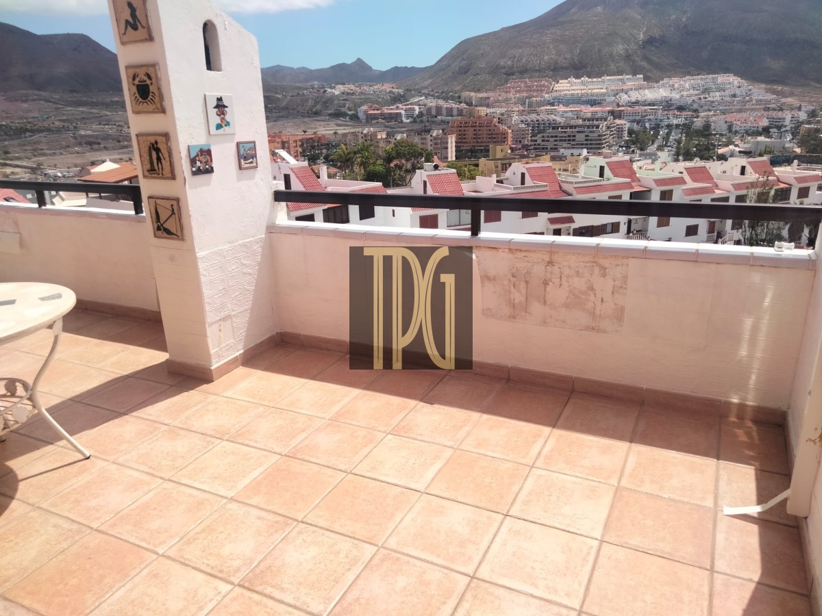 Terrace with tan tiled floor, white pillar with small tiles, and a round white table on the left, overlooking a town and mountains in the distance.