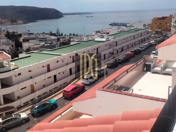 Balcony view over a coastal town: white apartment blocks with green roofs along a street, cars parked, blue sea beyond.
