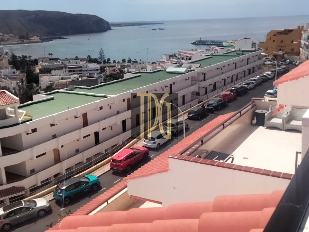 Balcony view over a coastal town: white apartment blocks with green roofs along a street, cars parked, blue sea beyond.