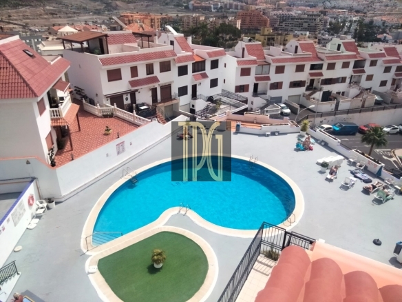Aerial view of a curved blue swimming pool with a pale deck, surrounded by sun loungers and white buildings with red-tiled roofs at a resort.