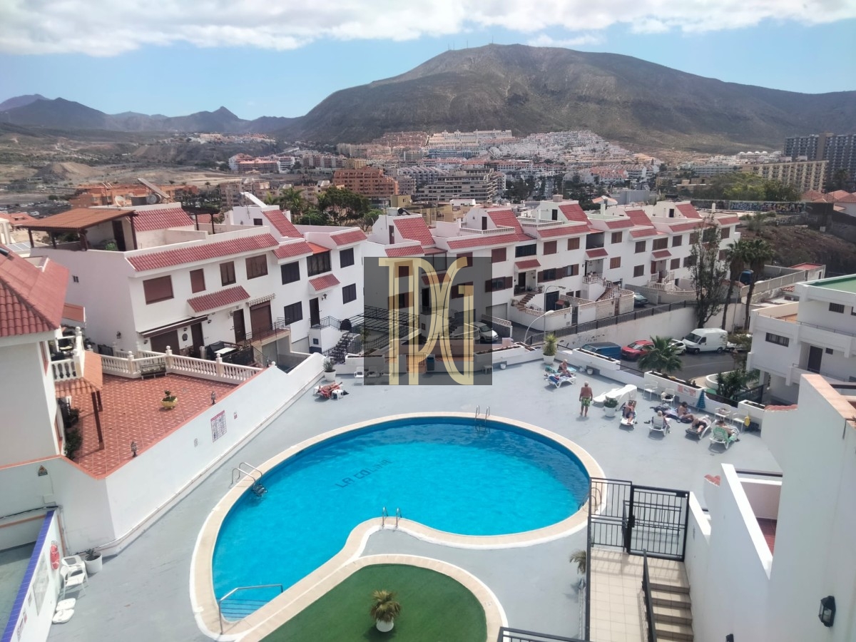 Aerial view of a resort pool area with a curved blue pool, surrounded by white buildings with red-tiled roofs and lounge chairs, with mountains in the distance.