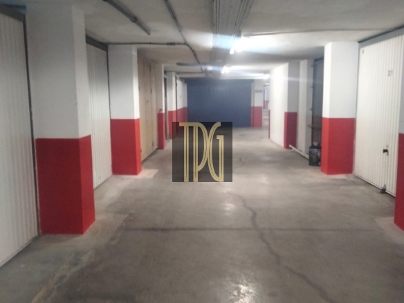 Underground parking garage hallway with white and red pillars, closed storage doors along both sides, and a long concrete corridor receding into the distance.
