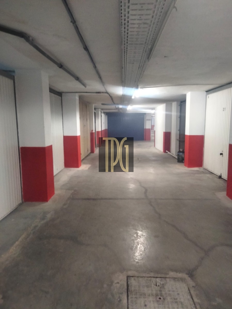 Underground parking garage hallway with white and red pillars, closed storage doors along both sides, and a long concrete corridor receding into the distance.