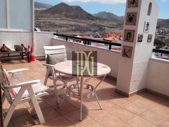 Sunny rooftop terrace with white patio furniture and a round table, overlooking hills and rooftops in a coastal town.