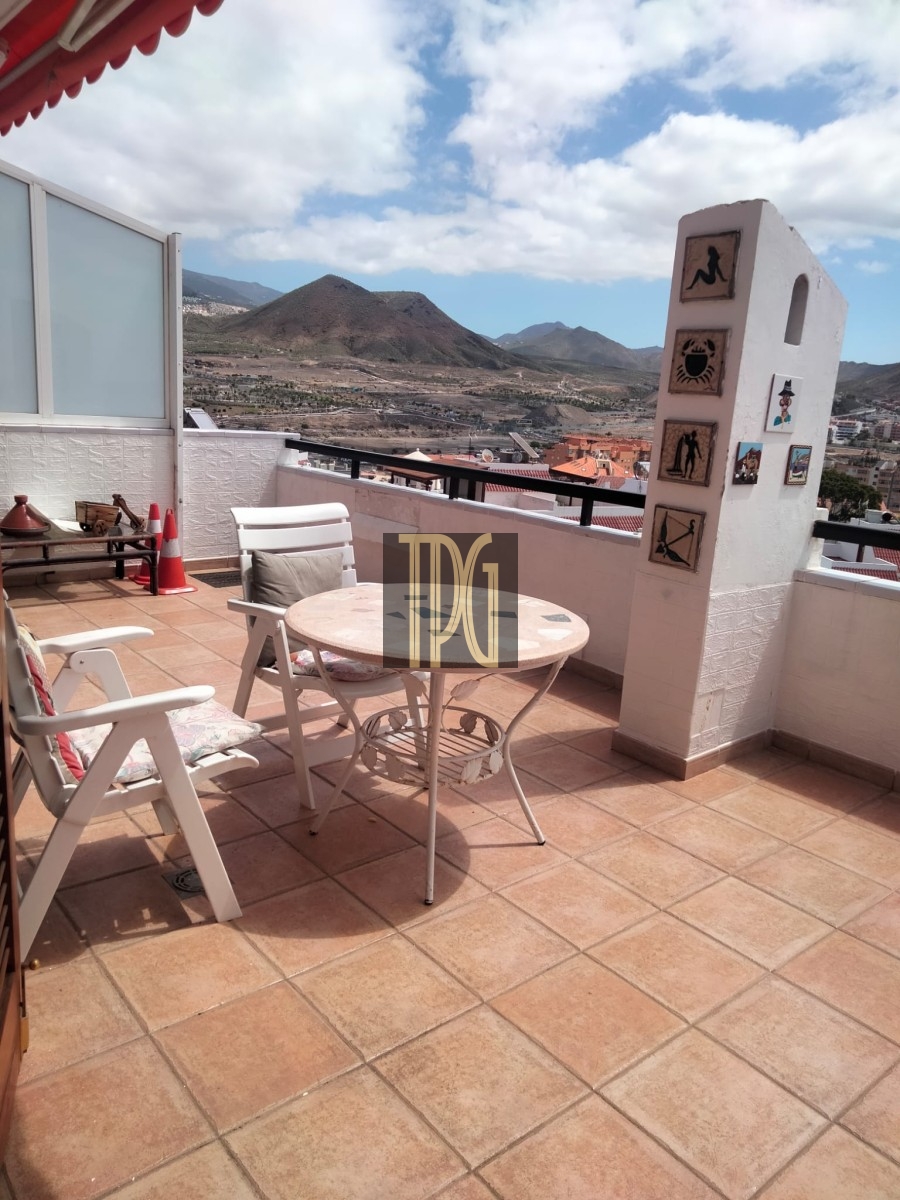 Sunny rooftop terrace with white patio furniture and a round table, overlooking hills and rooftops in a coastal town.