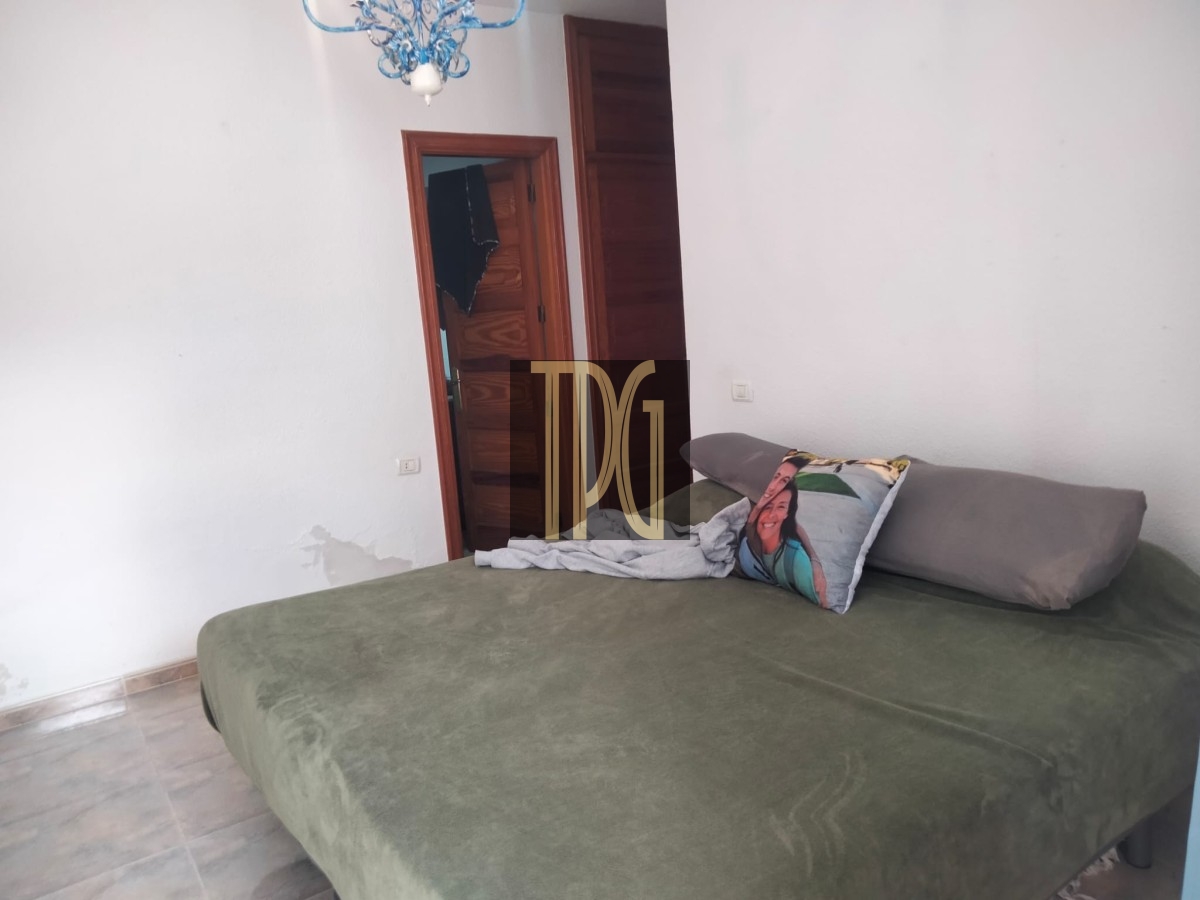 Bedroom with a green bedspread and gray pillows; a decorative pillow shows a person’s face, a white blanket folded on the bed, and a wooden door with a blue chandelier overhead.