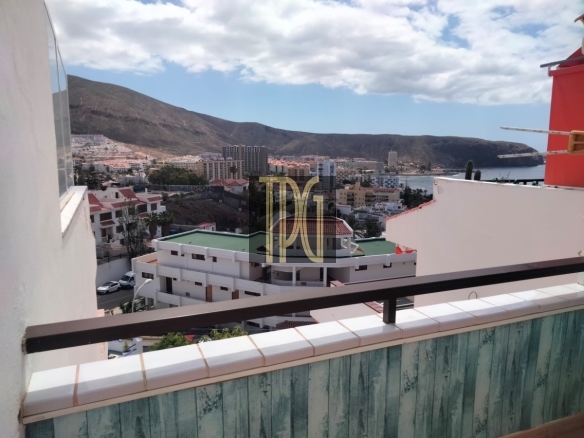 View from a balcony of a coastal city with white buildings, hills in the distance, and the sea under a partly cloudy sky.