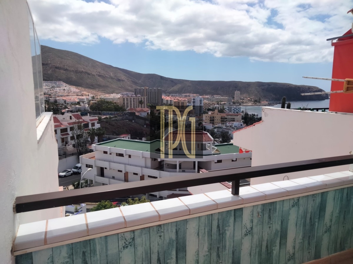 View from a balcony of a coastal city with white buildings, hills in the distance, and the sea under a partly cloudy sky.