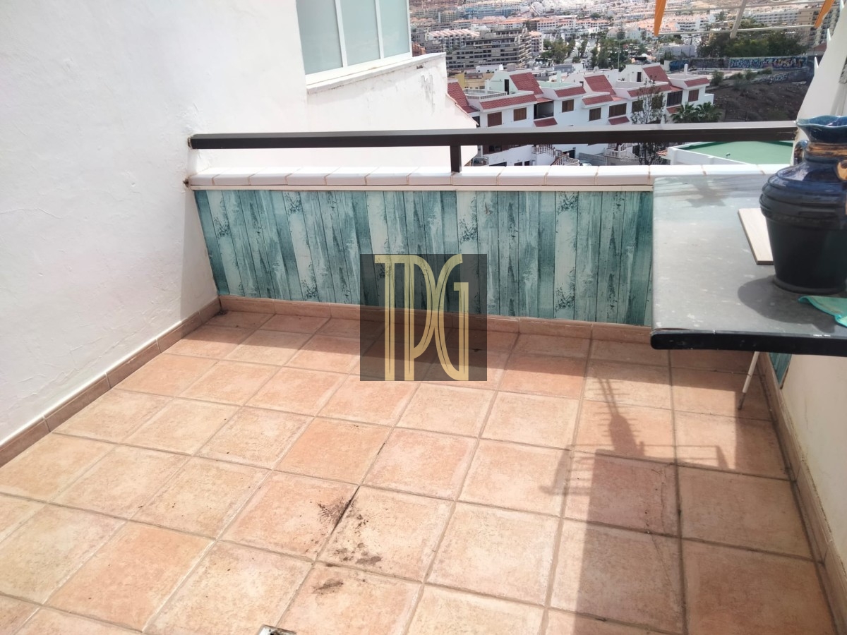 Sunny balcony with terracotta tiles, turquoise wooden paneling, and a black railing overlooking a cityscape; a table with a ceramic pot on the right.