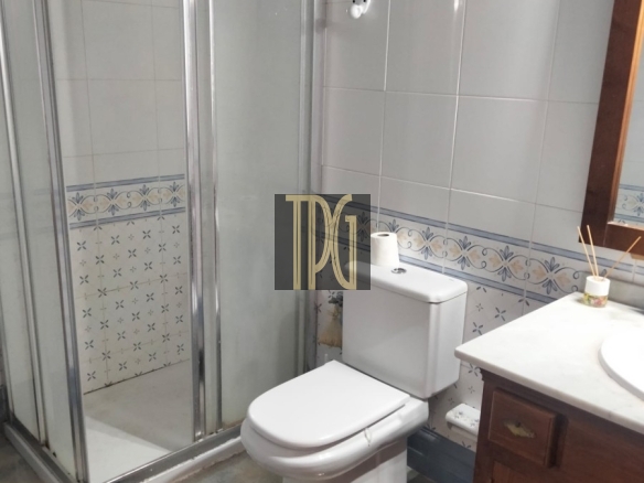 Small bathroom with a glass-enclosed shower, white toilet, and wooden vanity; blue tile border and a black bath mat on a tiled floor.