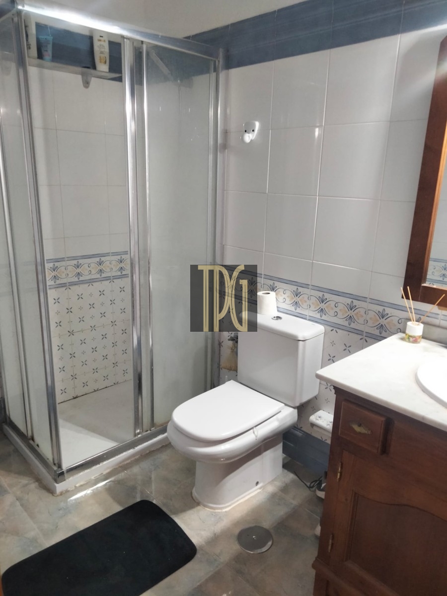 Small bathroom with a glass-enclosed shower, white toilet, and wooden vanity; blue tile border and a black bath mat on a tiled floor.