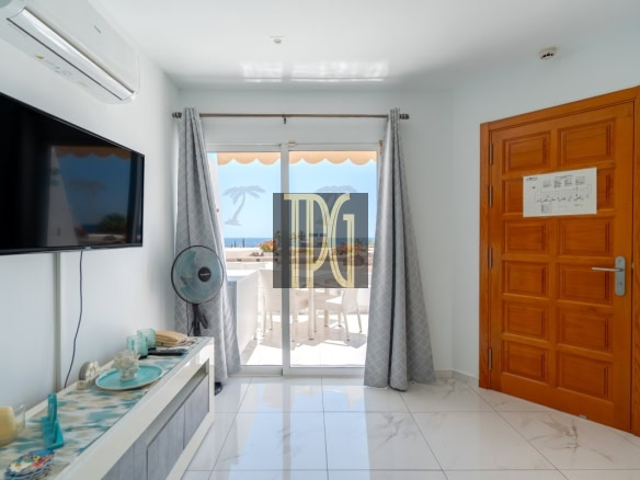 Living room with wall-mounted TV, ceiling AC, standing fan, and glass doors to a balcony with an ocean view.