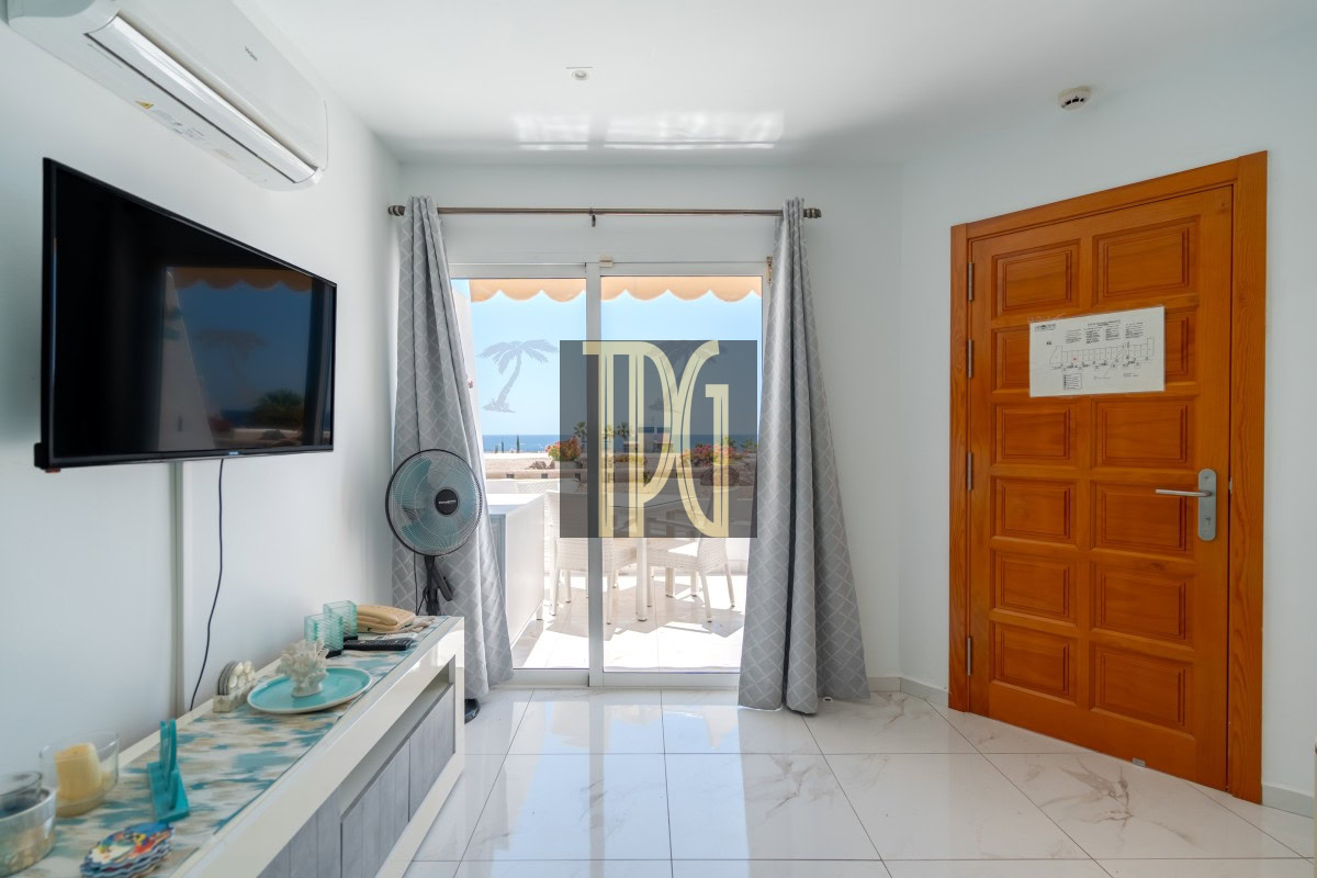Living room with wall-mounted TV, ceiling AC, standing fan, and glass doors to a balcony with an ocean view.