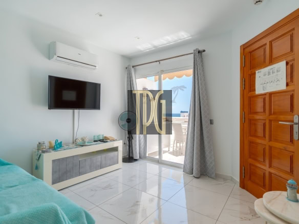 Living room with a wall-mounted TV, an air conditioner above, and a white media console facing a glass door to a balcony with ocean view.