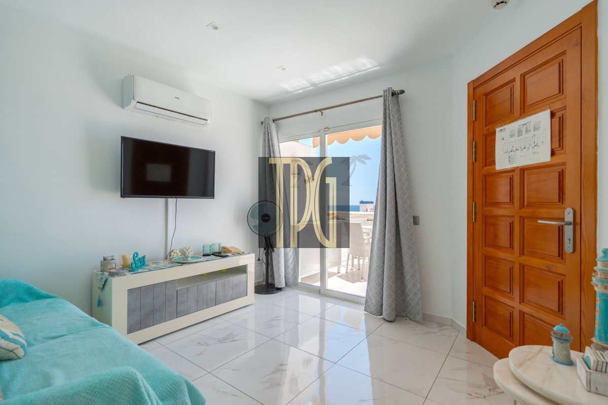 Living room with a wall-mounted TV, an air conditioner above, and a white media console facing a glass door to a balcony with ocean view.