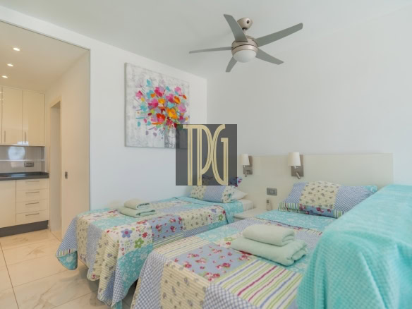 Twin beds with colorful patchwork quilts and folded towels in a bright hotel room; kitchenette visible through an open doorway and a ceiling fan overhead.